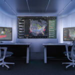 Interior view of a compact, modern control room with two workstations positioned on either side of the frame, each equipped with a desk, keyboard, mouse and large computer monitor. The monitors display complex data visualisations, including weather radar patterns on the left screen and dense network maps of flight paths and activity across North America on the central wall display and the right screen. Two ergonomic office chairs with mesh backs are positioned in front of the desks. The walls and ceiling are lined with smooth, light grey panels, and recessed lighting creates an evenly illuminated, high‑tech environment focused on monitoring and analysis.