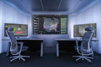 Interior view of a compact, modern control room with two workstations positioned on either side of the frame, each equipped with a desk, keyboard, mouse and large computer monitor. The monitors display complex data visualisations, including weather radar patterns on the left screen and dense network maps of flight paths and activity across North America on the central wall display and the right screen. Two ergonomic office chairs with mesh backs are positioned in front of the desks. The walls and ceiling are lined with smooth, light grey panels, and recessed lighting creates an evenly illuminated, high‑tech environment focused on monitoring and analysis.