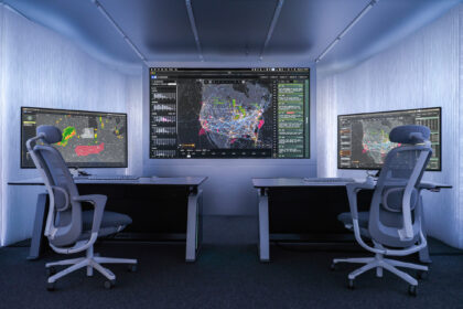 Interior view of a compact, modern control room with two workstations positioned on either side of the frame, each equipped with a desk, keyboard, mouse and large computer monitor. The monitors display complex data visualisations, including weather radar patterns on the left screen and dense network maps of flight paths and activity across North America on the central wall display and the right screen. Two ergonomic office chairs with mesh backs are positioned in front of the desks. The walls and ceiling are lined with smooth, light grey panels, and recessed lighting creates an evenly illuminated, high‑tech environment focused on monitoring and analysis.