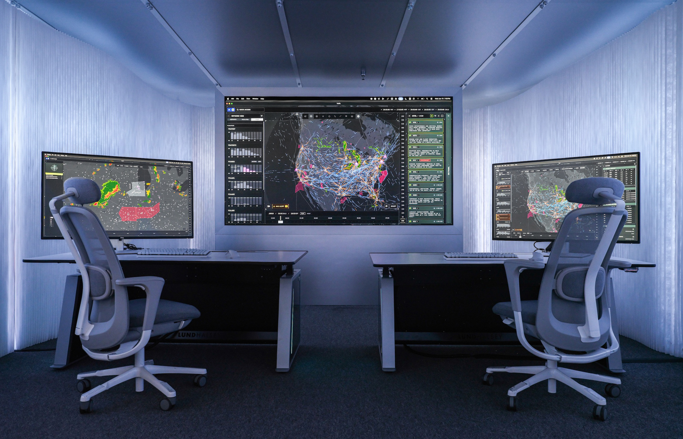 Interior view of a compact, modern control room with two workstations positioned on either side of the frame, each equipped with a desk, keyboard, mouse and large computer monitor. The monitors display complex data visualisations, including weather radar patterns on the left screen and dense network maps of flight paths and activity across North America on the central wall display and the right screen. Two ergonomic office chairs with mesh backs are positioned in front of the desks. The walls and ceiling are lined with smooth, light grey panels, and recessed lighting creates an evenly illuminated, high‑tech environment focused on monitoring and analysis.