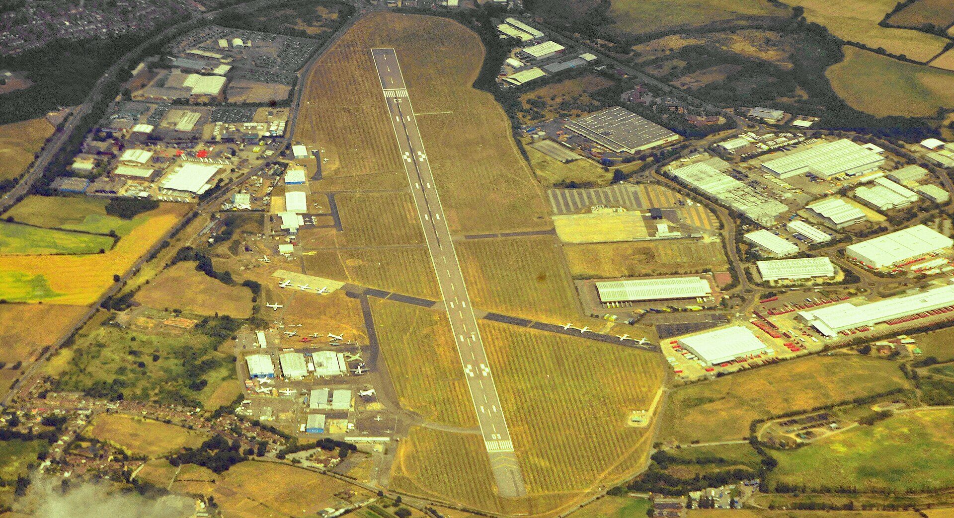 An aerial shot of Coventry Airport in 2010. The buildings are in view and in the middle is a long runway. Housing estates and fields are in the surroundings.