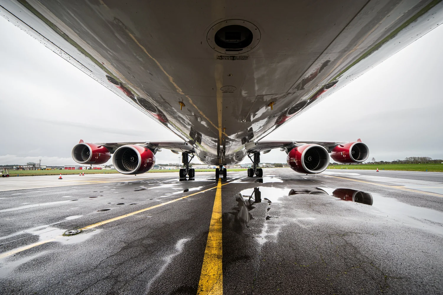 A low-angle view beneath a large four-engine aircraft shows the underside of the fuselage and landing gear positioned on a wet airport apron, with four red engines visible on either side and reflections in puddles along a yellow centerline under overcast skies.