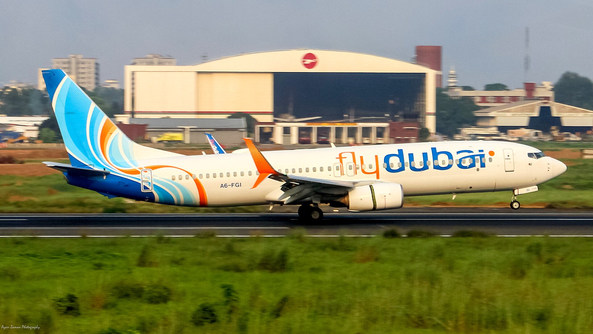 Flydubai Boeing 737-8 aircraft taking off from the runway toward the right side. Grey sky seen in the background, with a beige building. Grass in the foreground.
