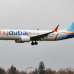 A Flydubai Boeing 737-8 MAX aircraft flying, coming in to land with its landing gear out. Trees seen underneath the aircraft and a grey sky in the background.