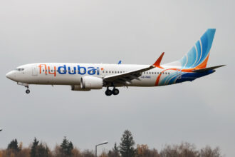 A Flydubai Boeing 737-8 MAX aircraft flying, coming in to land with its landing gear out. Trees seen underneath the aircraft and a grey sky in the background.