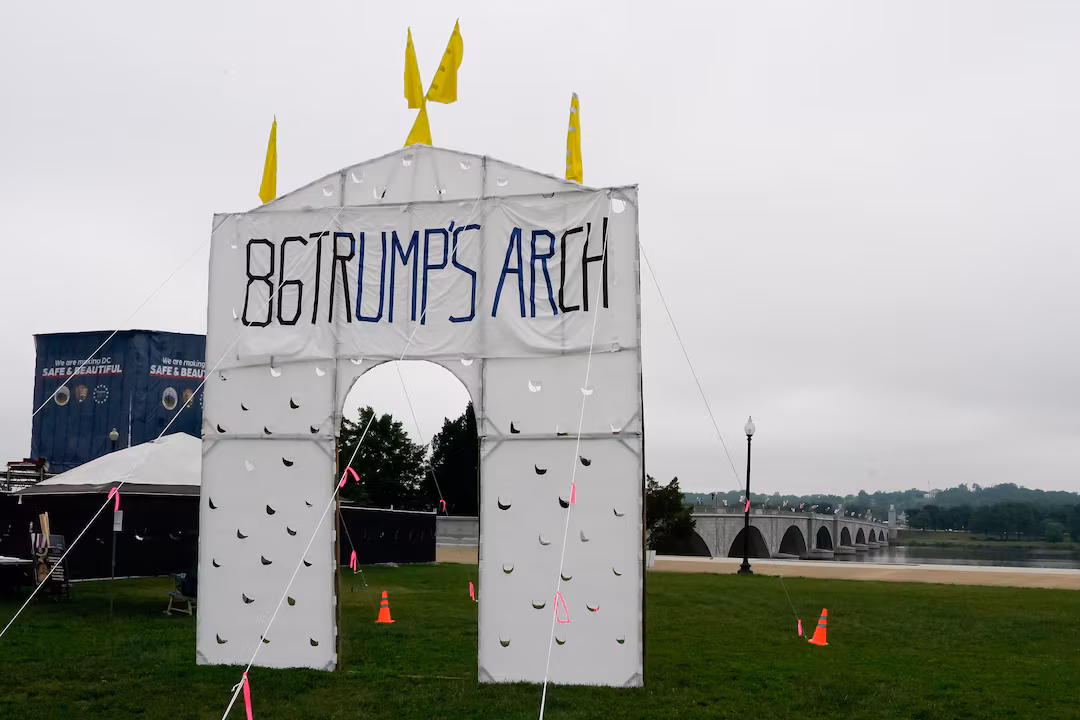 A mock arch stands erected on green grass with Arlington Memorial Bridge visible in the background. It appears to be made of some type of white sheeting, yellow flags, and metal bars. '86 TRUMP'S ARCH' is written on the front. The sky is grey and cloudy in the background. 
