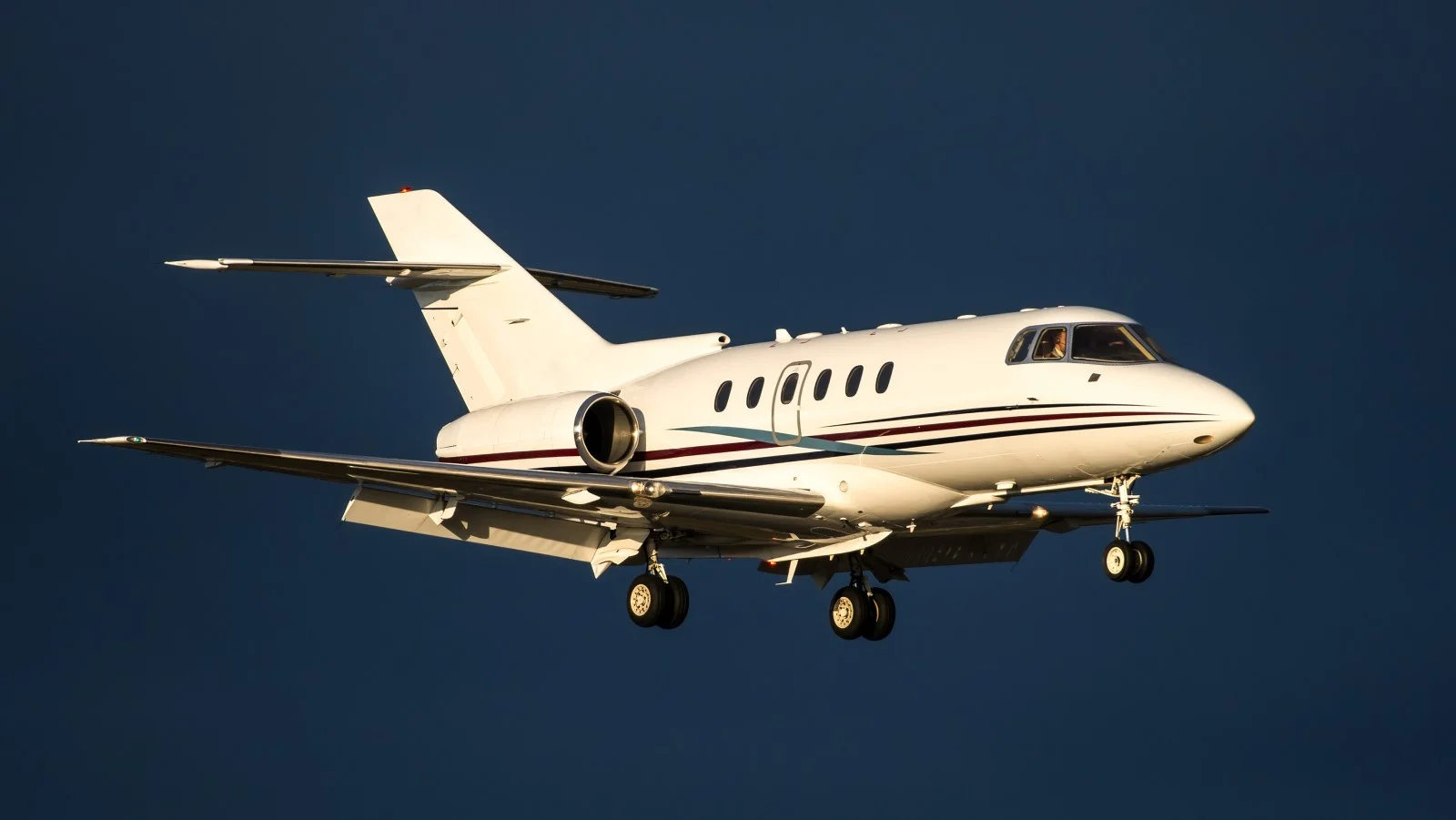 A Hawker 800XP plane mid-flight in front of a dark blue sky. The landing gear is out.