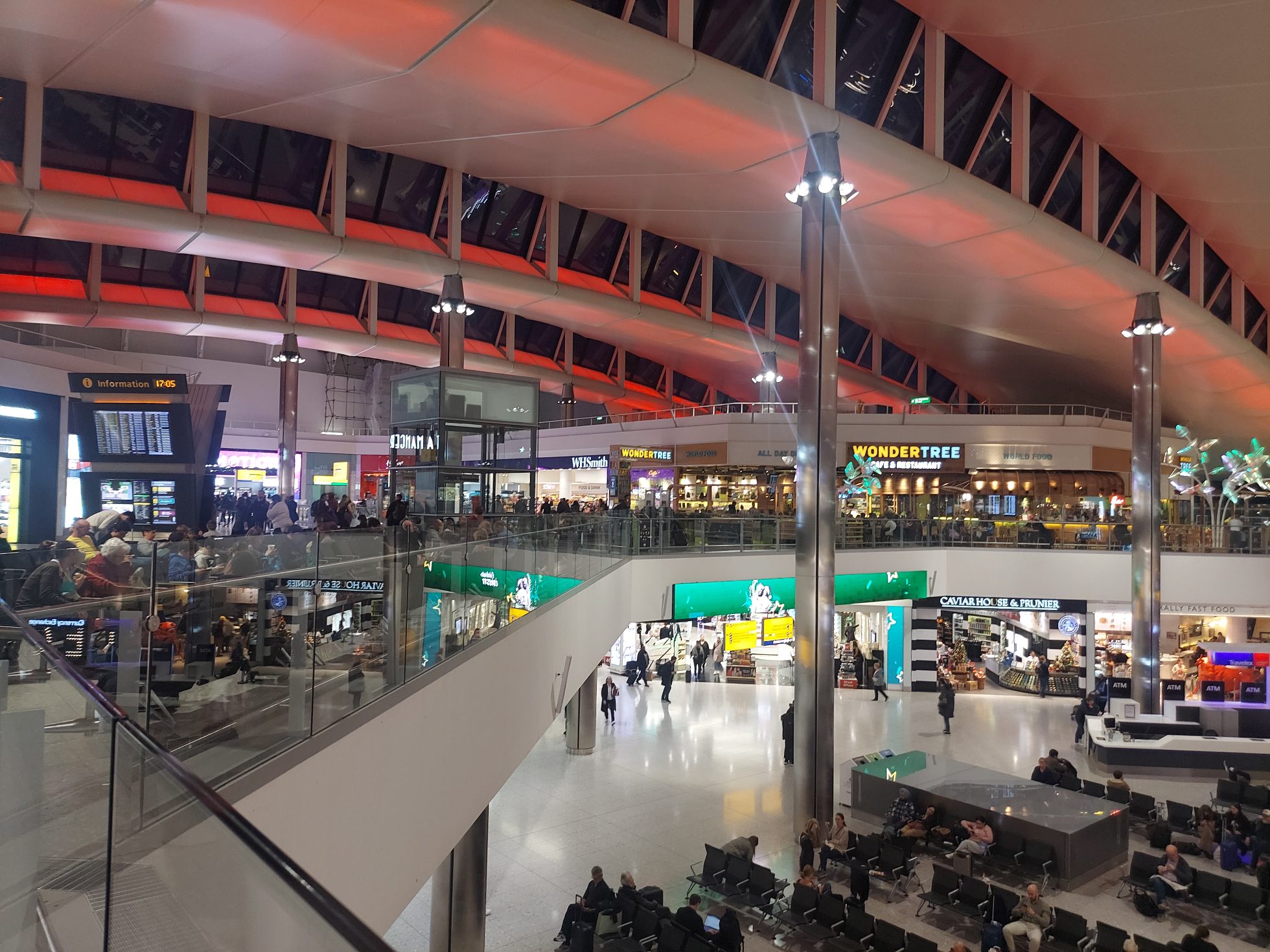 The view from a bridge in the departure lounge of London Heathrow Airport. The lounge is full of people walking in and out of various cafes and shops.