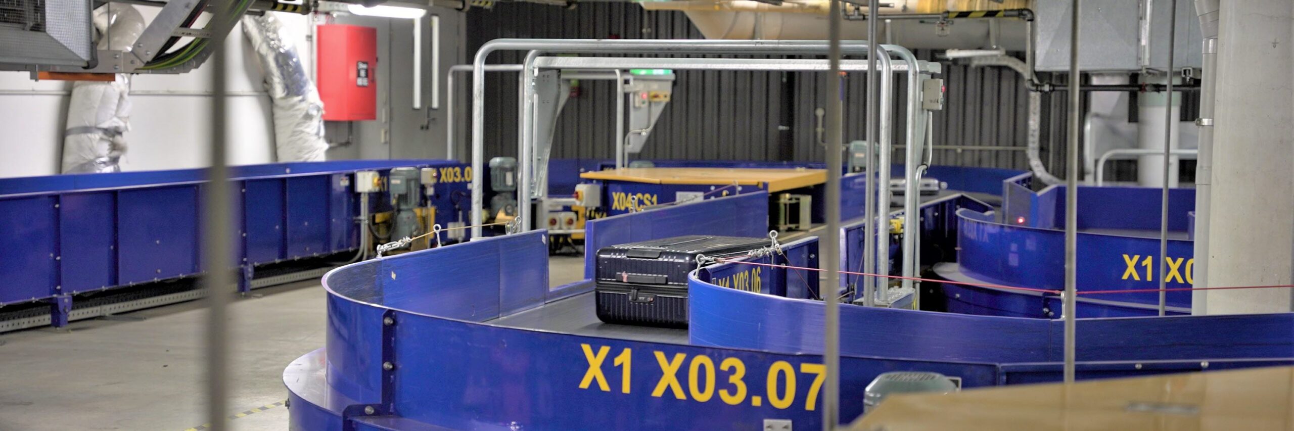 A single black suitcase on an airport baggage conveyor belt. The walls of the belt are blue with yellow writing.