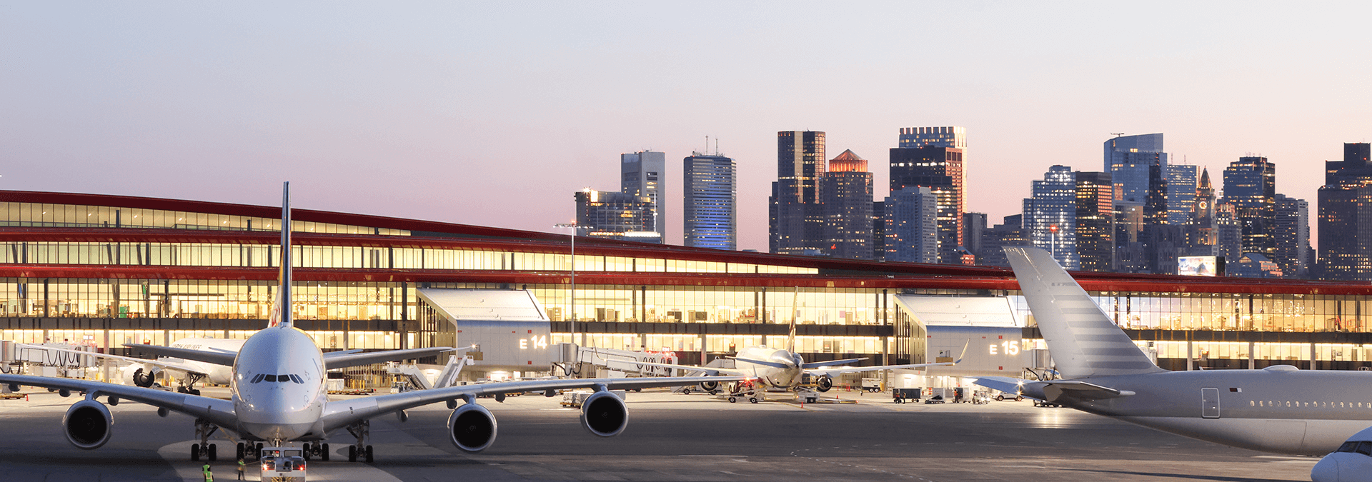 BOS airport at sunset, showing a white jet in front of a well-lit terminal building, and the Boston skyline behind.