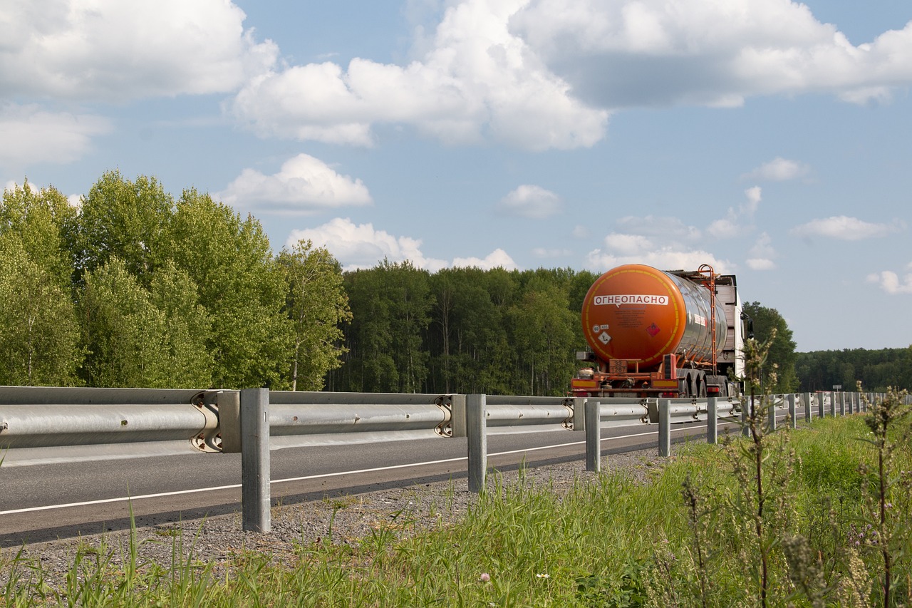 An orange oil truck drives down a road next to a grassy verge and a green forest, in front of a cloudy sky.