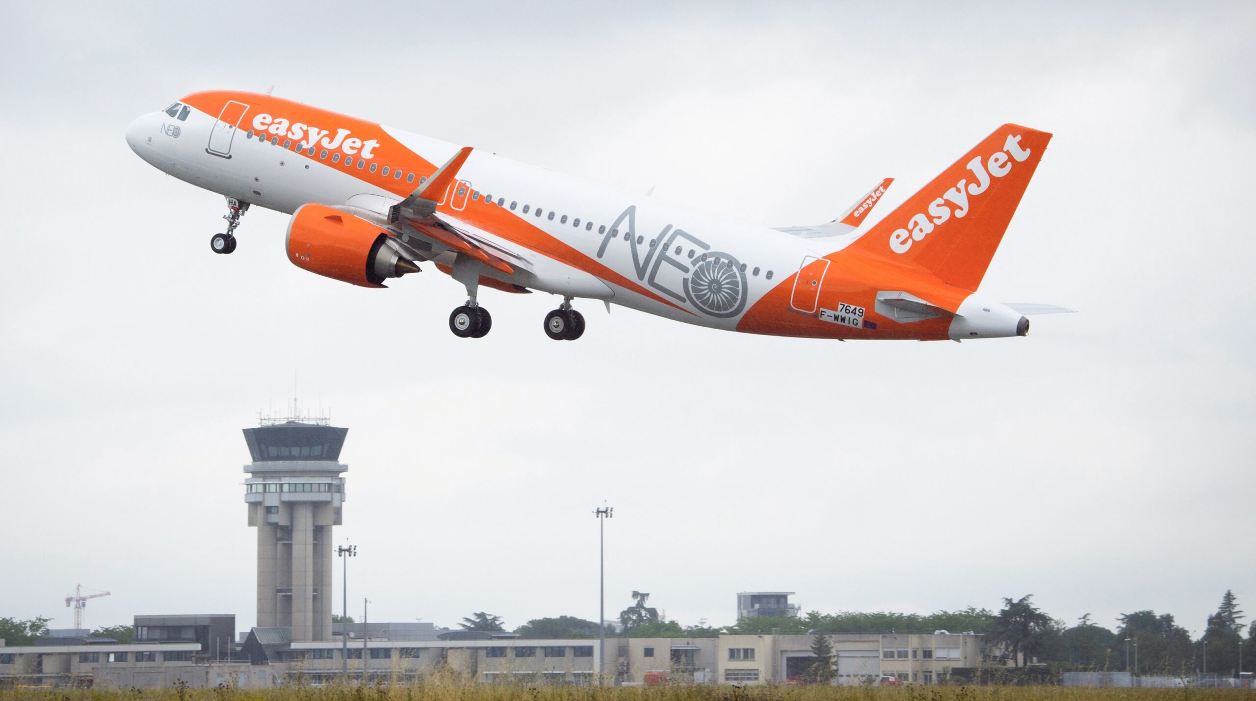 An EasyJet Airbus A320neo flying upwards over an airport terminal.