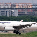 A China Eastern Airbus A330 jet descending towards a runway with a grassy verge and a city skyline behind.