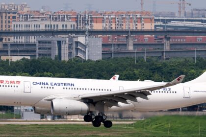 A China Eastern Airbus A330 jet descending towards a runway with a grassy verge and a city skyline behind.