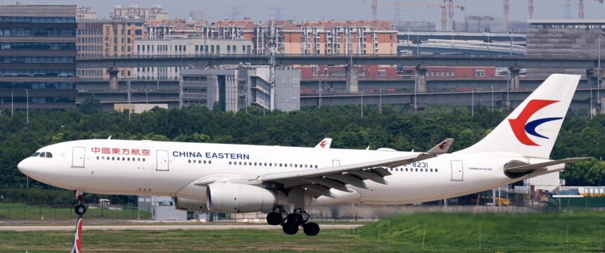 A China Eastern Airbus A330 jet descending towards a runway with a grassy verge and a city skyline behind.