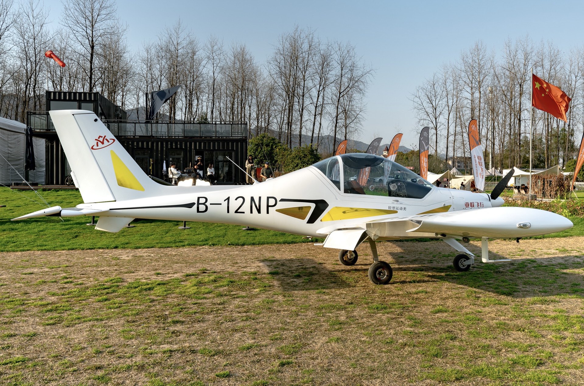 A yellow and white Sunward Aurora SA60L plane parked on a grass field in front of a Chinese national flag.