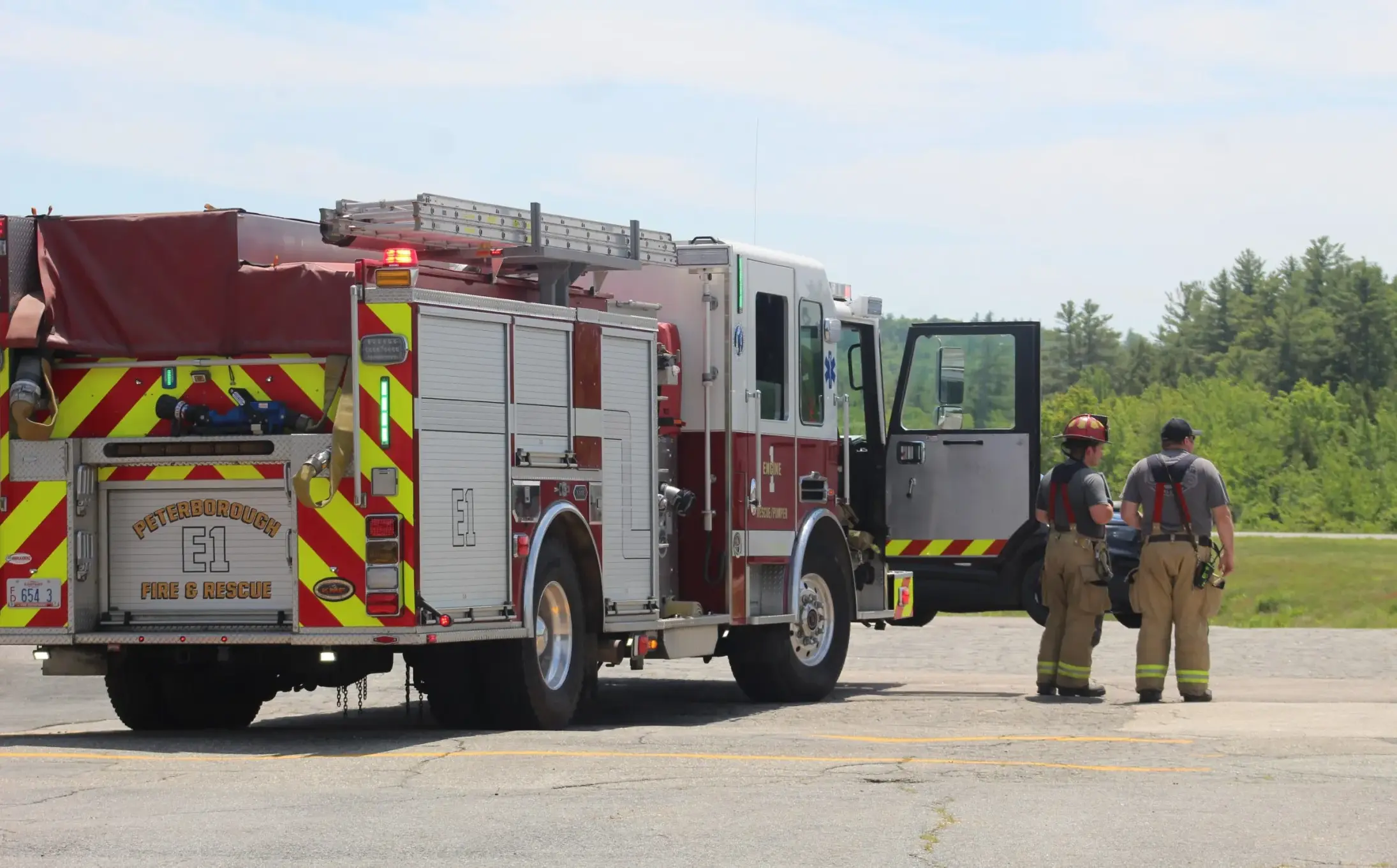 A fire truck on the left side with the door open. Two male firefighters stand beside it on the right looking out into the field. Blue sky and trees in the background.