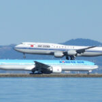 Side view of two large commercial aircraft at an airport beside a body of water. In the foreground, a light blue and white Korean Air Boeing 777 taxis along the runway, with “KOREAN AIR” written along the fuselage and the airline’s red-and-blue yin‑yang logo on the tail. Behind it, an Air China Boeing 747 in white livery with a blue stripe and red phoenix logo on the tail descends to land, its landing gear extended and positioned just above the Korean Air aircraft. Calm water occupies the lower foreground, while low hills and a hazy skyline form the background under a pale blue sky.