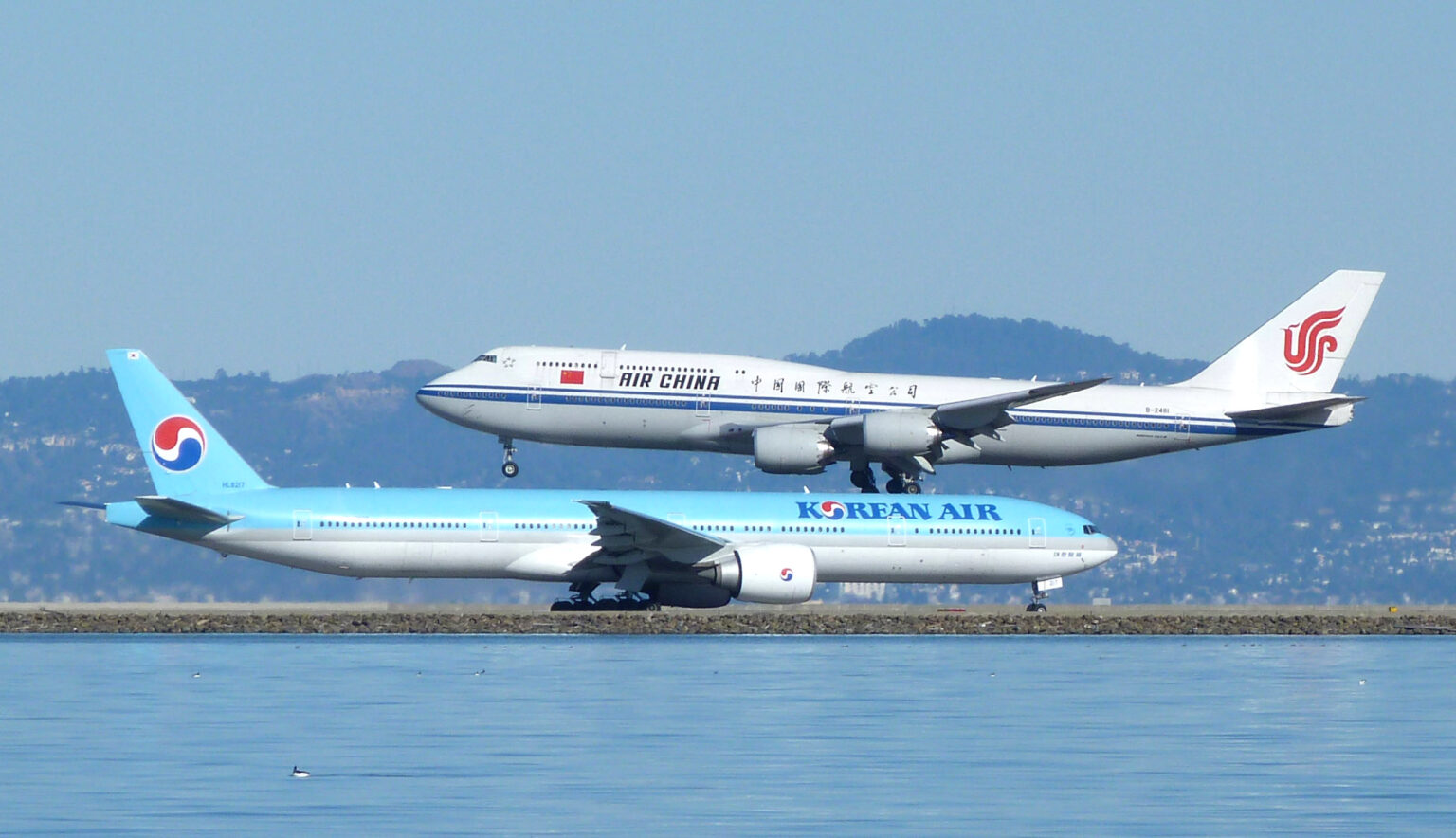 Side view of two large commercial aircraft at an airport beside a body of water. In the foreground, a light blue and white Korean Air Boeing 777 taxis along the runway, with “KOREAN AIR” written along the fuselage and the airline’s red-and-blue yin‑yang logo on the tail. Behind it, an Air China Boeing 747 in white livery with a blue stripe and red phoenix logo on the tail descends to land, its landing gear extended and positioned just above the Korean Air aircraft. Calm water occupies the lower foreground, while low hills and a hazy skyline form the background under a pale blue sky.