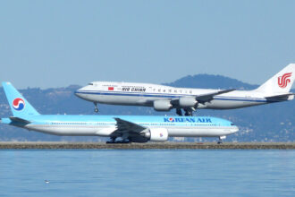 Side view of two large commercial aircraft at an airport beside a body of water. In the foreground, a light blue and white Korean Air Boeing 777 taxis along the runway, with “KOREAN AIR” written along the fuselage and the airline’s red-and-blue yin‑yang logo on the tail. Behind it, an Air China Boeing 747 in white livery with a blue stripe and red phoenix logo on the tail descends to land, its landing gear extended and positioned just above the Korean Air aircraft. Calm water occupies the lower foreground, while low hills and a hazy skyline form the background under a pale blue sky.