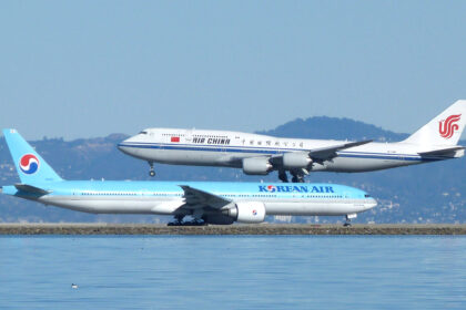 Side view of two large commercial aircraft at an airport beside a body of water. In the foreground, a light blue and white Korean Air Boeing 777 taxis along the runway, with “KOREAN AIR” written along the fuselage and the airline’s red-and-blue yin‑yang logo on the tail. Behind it, an Air China Boeing 747 in white livery with a blue stripe and red phoenix logo on the tail descends to land, its landing gear extended and positioned just above the Korean Air aircraft. Calm water occupies the lower foreground, while low hills and a hazy skyline form the background under a pale blue sky.