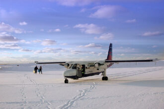 This image shows a Britten-Norman BN-2 Islander aircraft operated by Loganair (sporting a British Airways Express livery) sitting on the snow-covered Fair Isle airstrip in Shetland, Scotland.
