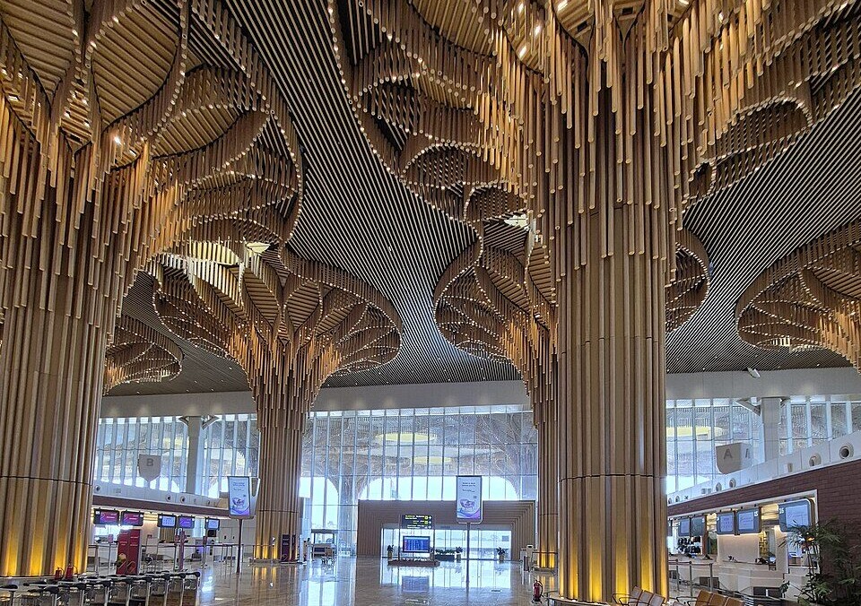 The main atrium can be seen. Large columns made from what looks to be bamboo extend in a distinctive tree like shape at equal intervals. Rows of seats are positioned by a column in the centre right of the image, and bag trolly storage can be seen opposite. Various help desks can be seen vaguely in the background on the left and right, and in the centre background the entire back wall is glass.