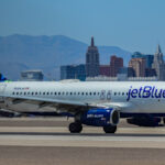Side view of a JetBlue Airways Airbus A320 taxiing on a runway, painted in a white and dark blue livery with “jetBlue” written along the fuselage. The aircraft’s engines are deep blue with “jetblue.com” printed on them, and the tail features a mosaic pattern in shades of blue and green. The landing gear is fully extended as it rolls along the tarmac. In the background, a city skyline of tall hotels and distinctive high‑rise buildings stands against hazy mountains under a clear blue sky, suggesting an airport near an urban, desert landscape.