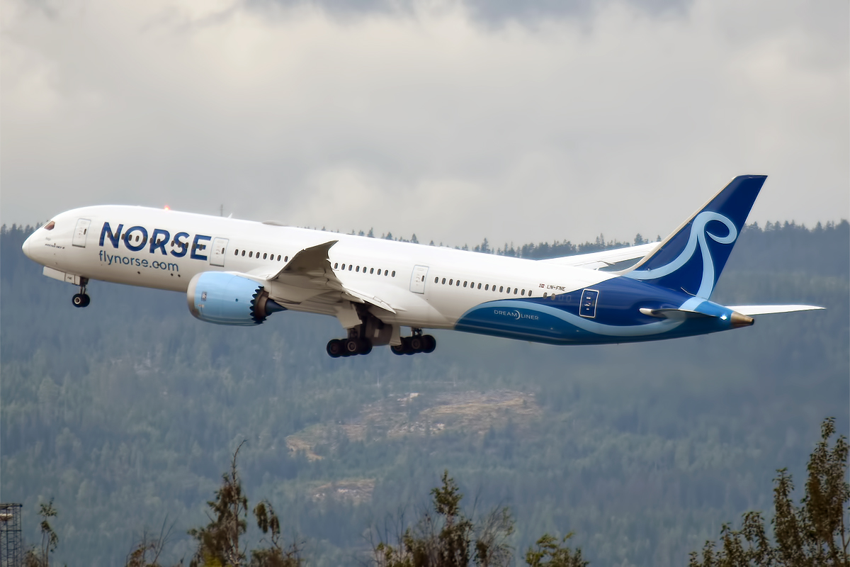 A Norse Atlantic aircraft mi-flight. The plane is mostly white with mid-blue branding that reads 'NORSE' close to front door, and a mid-blue tail with a light blue logo. The engines are also light blue. The nose of the aircraft is on the left of the image, at an upwards angle facing away from the camera, while the tail is on the lower centre-right - the aircraft is ascending away from the camera. Mountainous forests can be seen in the lower background of the image, while cloudy sky can be seen in the upper background. 