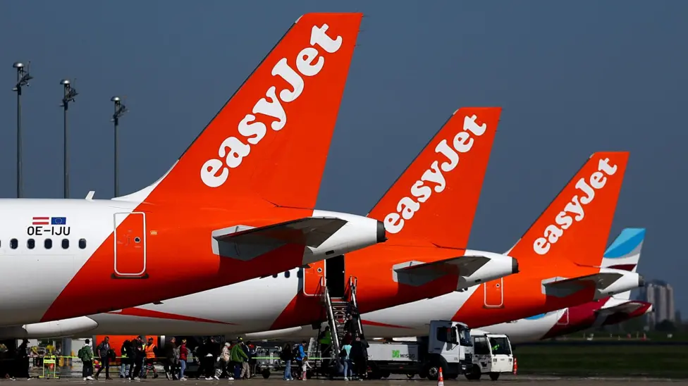 EasyJet aircraft lined up at a terminal whilst passengers begin to board. The rear wing of the plane displays the airline's "iconic" orange colour.