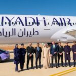 A Riyadh Air B787 on the ground under a blue sky. The aircraft's livery is mostly white with purple accents. In front of the plane, one can see a car with the same colours as the aircraft and a group of nine people posing for the picture.