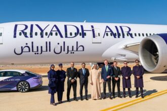 A Riyadh Air B787 on the ground under a blue sky. The aircraft's livery is mostly white with purple accents. In front of the plane, one can see a car with the same colours as the aircraft and a group of nine people posing for the picture.