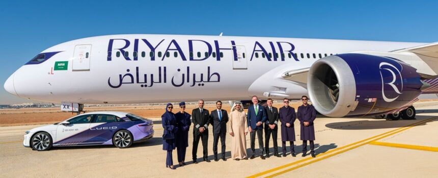 A Riyadh Air B787 on the ground under a blue sky. The aircraft's livery is mostly white with purple accents. In front of the plane, one can see a car with the same colours as the aircraft and a group of nine people posing for the picture.