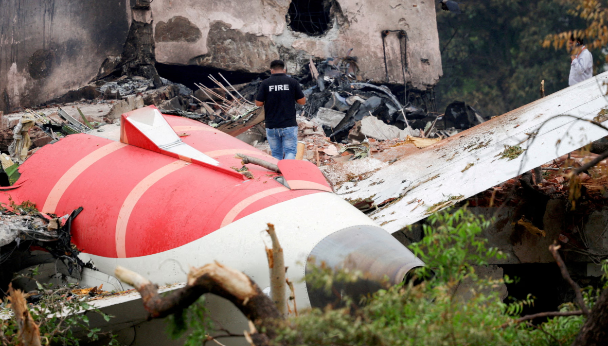 The wreckage of a small red and white plane lying in the trees with a man standing next to it and concrete in the background.