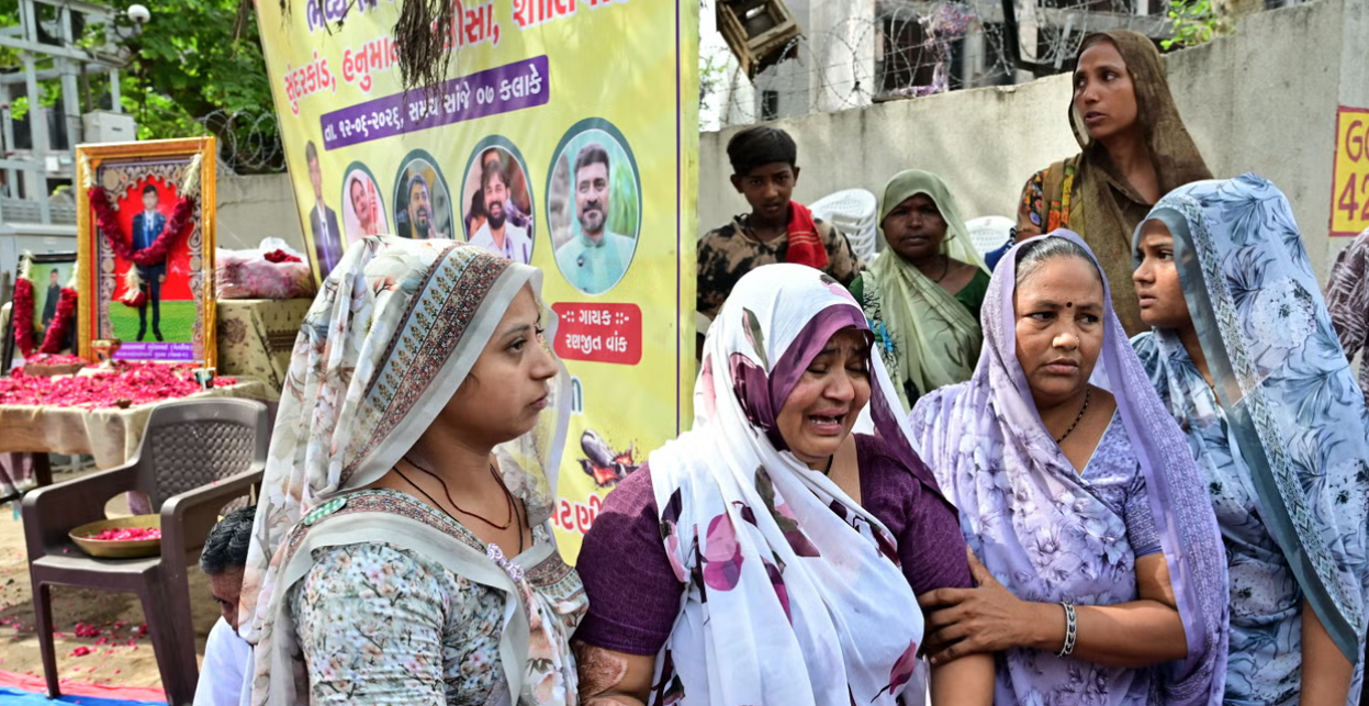A group of women dressed in traditional saris stands in front of a large poster and a framed photograph of a boy.