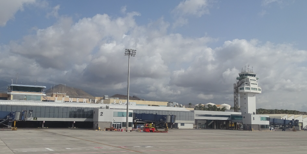 A grey and white airport terminal with tinted windows beside an air traffic control tower with mountains in the background and a cloudy sky.