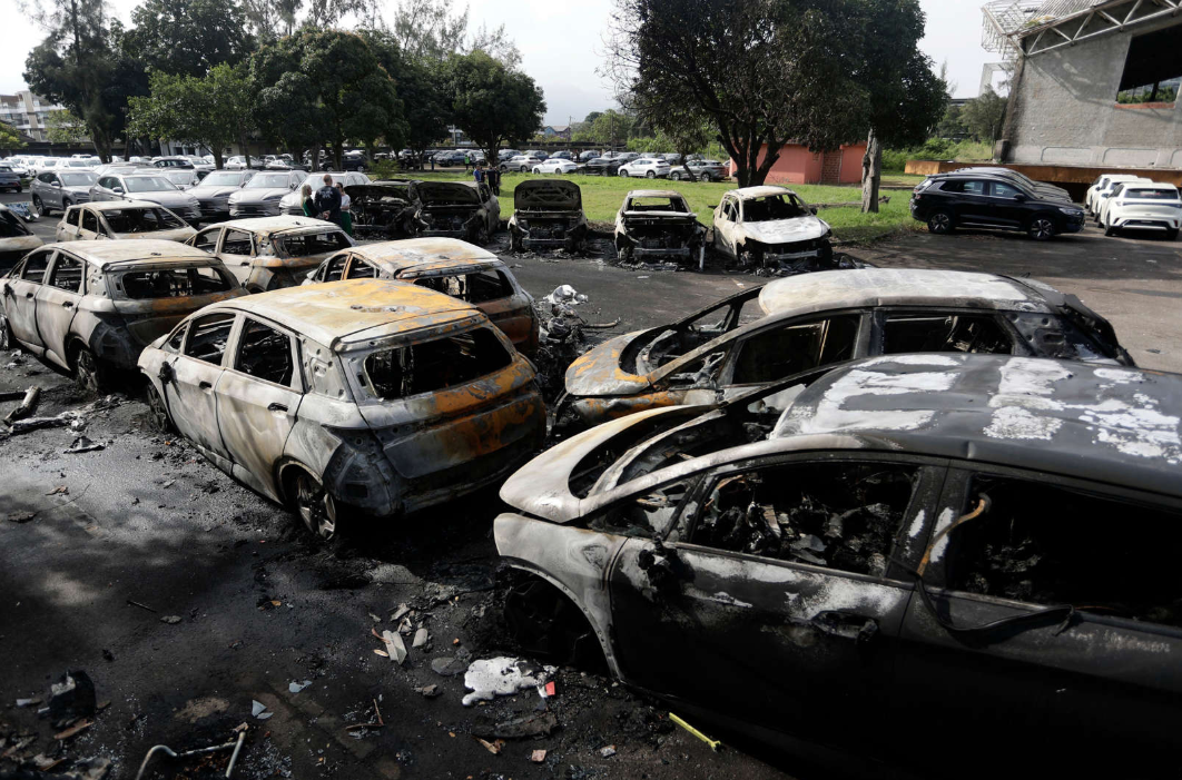 A street lined with damaged and burnt out cars with trees and grass in the background