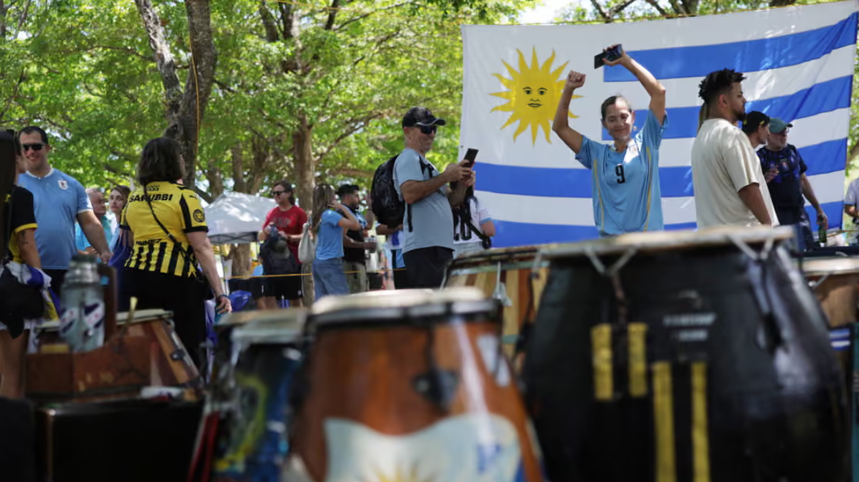 A group of people standing behind a set of drums with trees and the blue and white Uruguay flag in the background