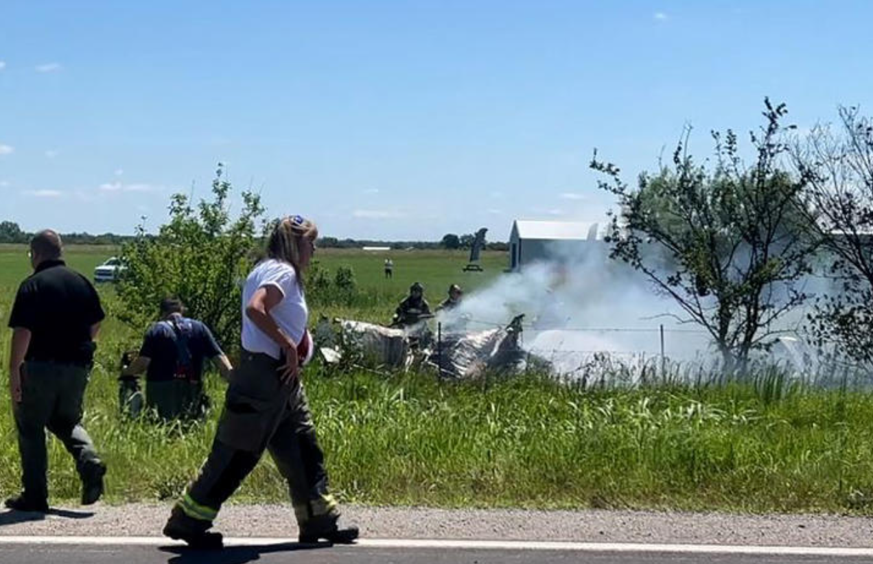 People walking on a country road with the wreckage of a plane behind with smoke coming out of it
