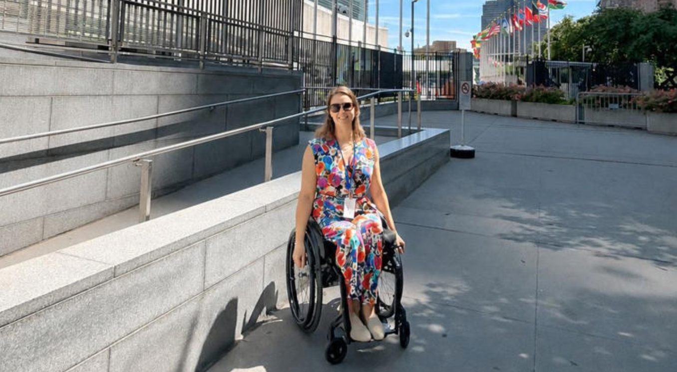 A woman wearing a floral dress and sitting in a wheelchair with a large grey building and flags in the background