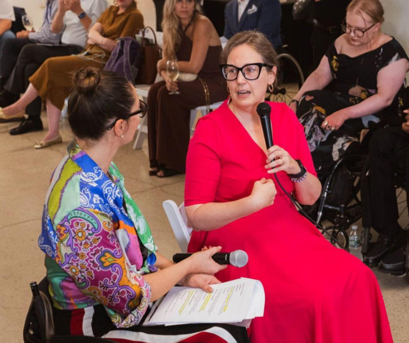 Two women sitting down and talking with microphones, with people watching in the background