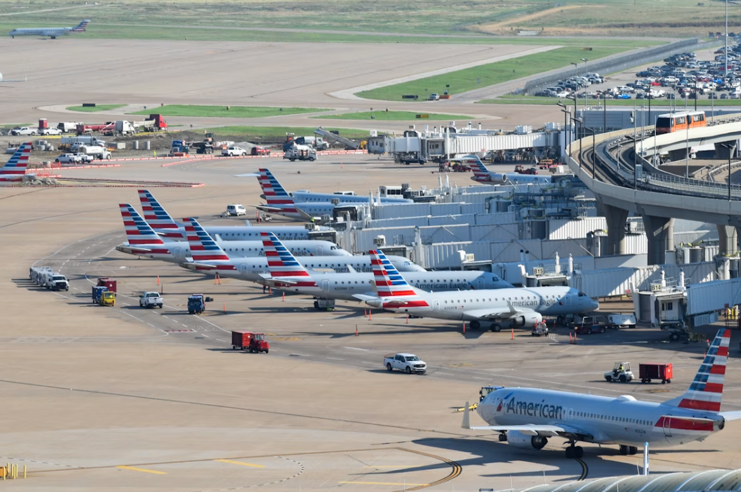 An airport filled with lots of airplanes parked on top of tarmac, with a runway and grass in the background.