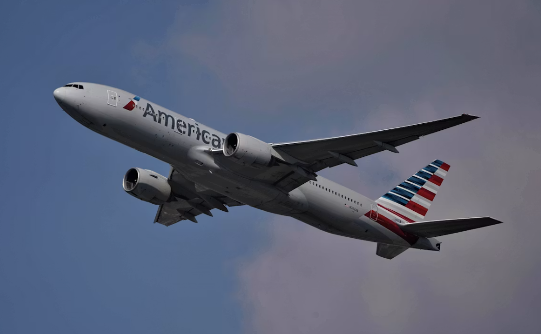A large white jetliner with the American flag flying through a cloudy sky.