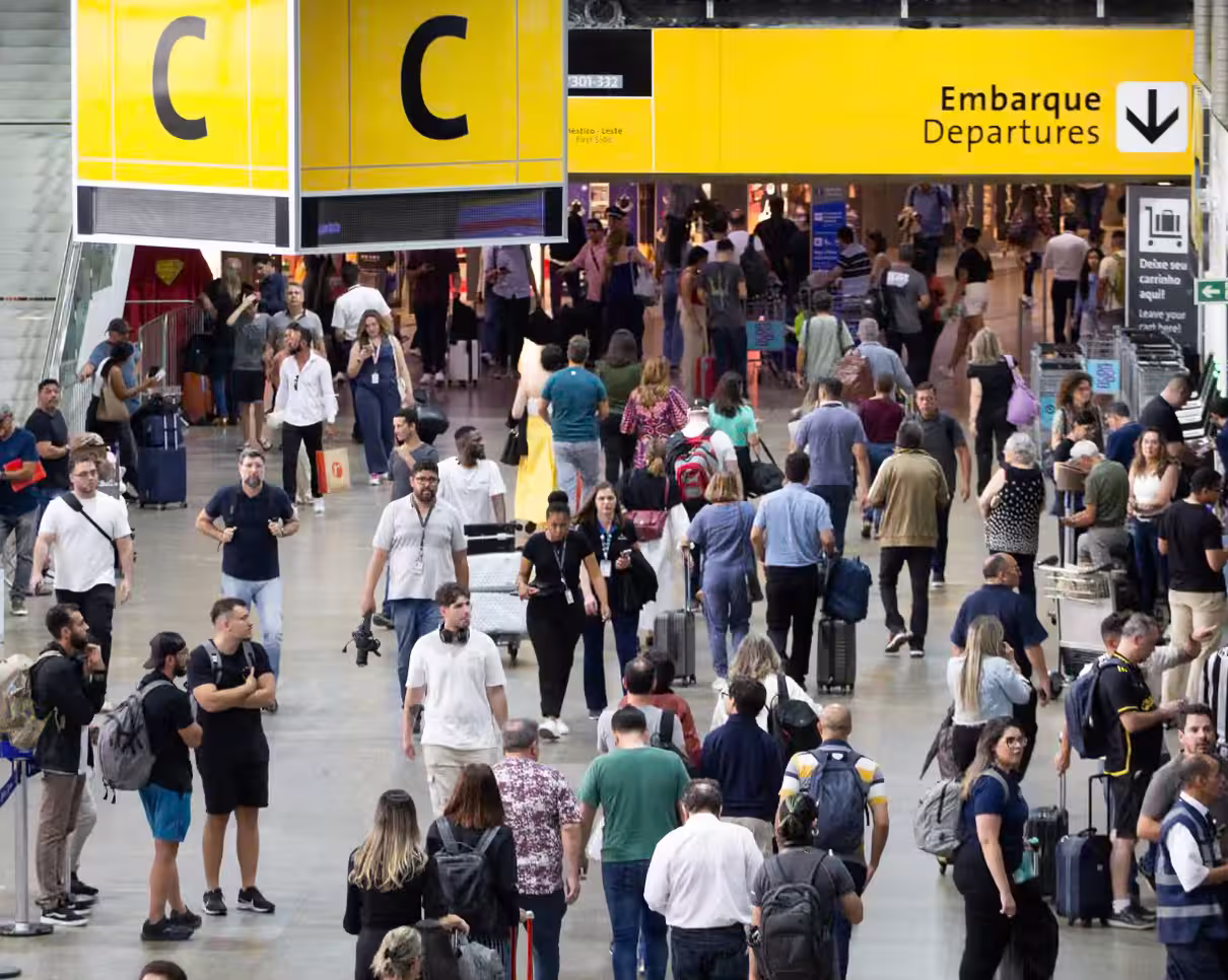 Several travellers seen at São Paulo International Airport (GRU). The crowd is seen above a grey floor and under two large yellow signs. 