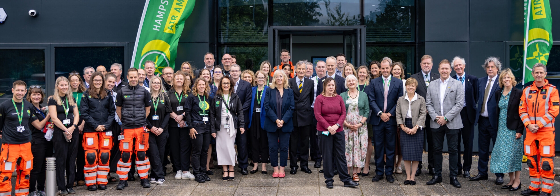 Image shows a large group of people including Air Ambulance staff wearing orange standing outside the new Hampshire & Isle of Wight Air Ambulance Headquarters at Southampton Airport