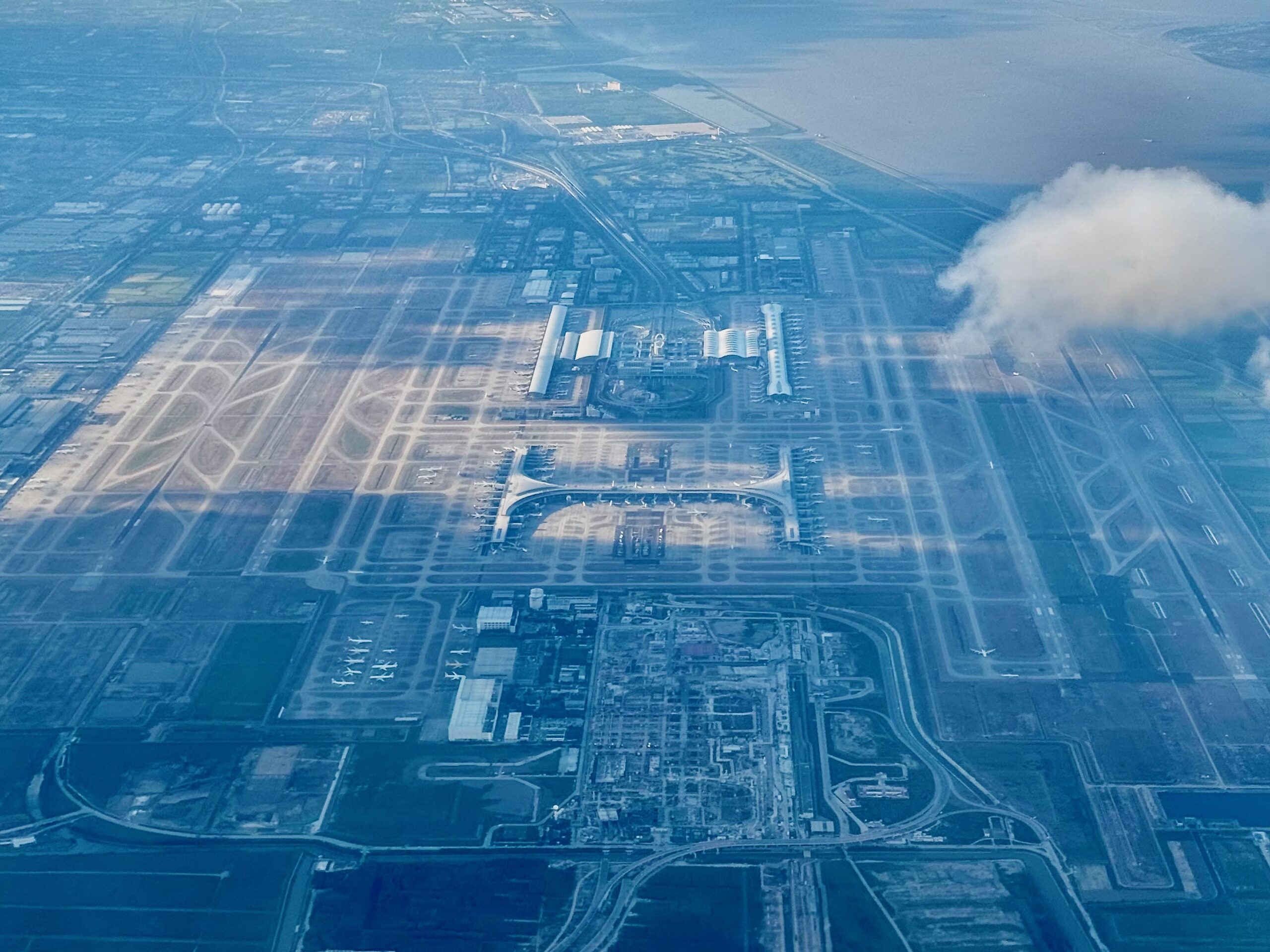 High, aerial view of a large coastal airport complex seen from above, with multiple runways and taxiways laid out in a grid across reclaimed land beside the sea. In the centre, a modern terminal building with curved roof sections is surrounded by aircraft stands, where several aeroplanes are parked. Long runways stretch parallel to the shoreline on the right, bordered by water and a narrow strip of land, while roads, service buildings and industrial facilities fill the surrounding area. A thin layer of haze and a patch of cloud partially obscure the scene, giving it a soft blue tint under daylight.