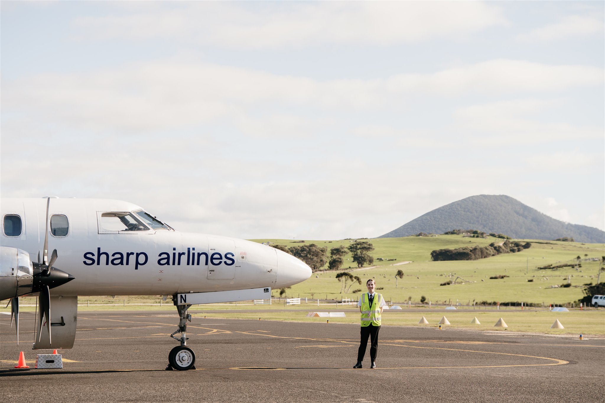 Half of a Sharp Airline's aircraft is seen in the picture. A worker wearing a neon yellow safety vest is standing in front of the aircraft posing for the picture. The background consists of a green mountain and a very light blue sky with clouds. 