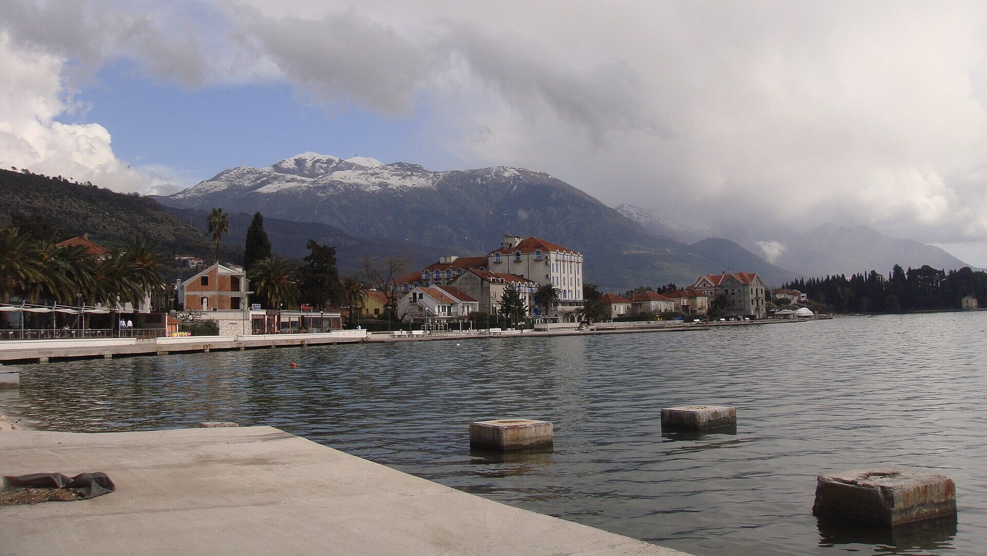 Wide-shot view of Tivat, Montenegro. Mountain in the background, with buildings in front of it. A lake in the foreground, surrounded by trees. A blue cloudy sky above.