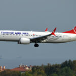 A Turkish Airlines B737-8 MAX flying over Tallinn, Estonia. The sky is grey and the plane is mostly white, with blue and red accents. Under the plane there are several brick ceilings and tree tops.