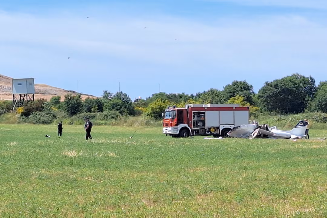 Small aircraft crash site. Police and firefighters attend the scene. Fire truck seen in the background behind the crash. Blue sky and trees in the background.